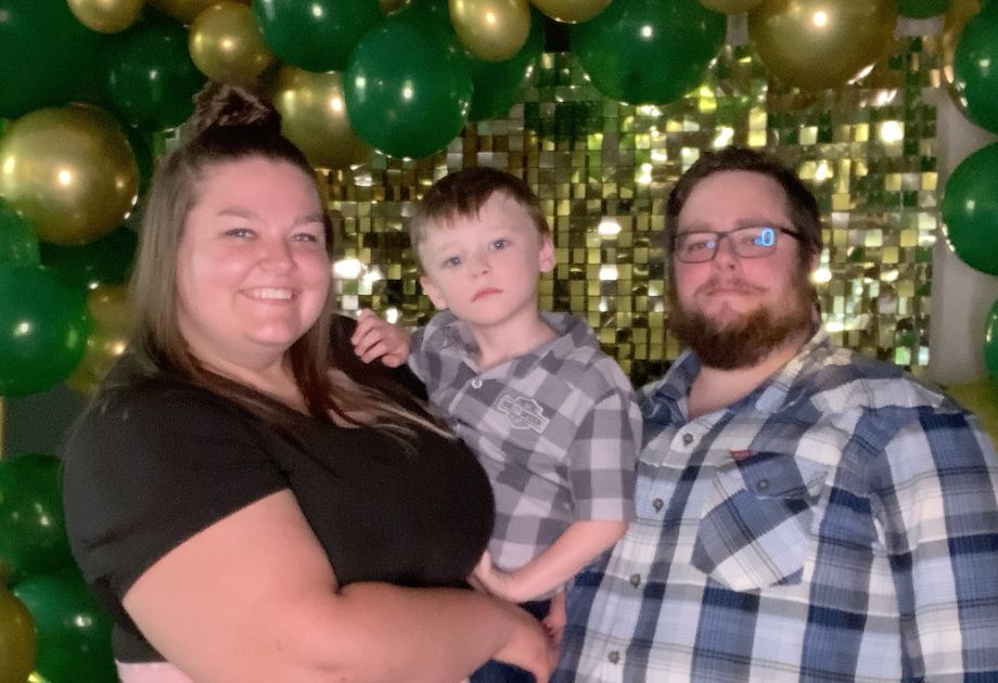 A smiling woman holding a young boy stands next to a man in front of an arch of balloons